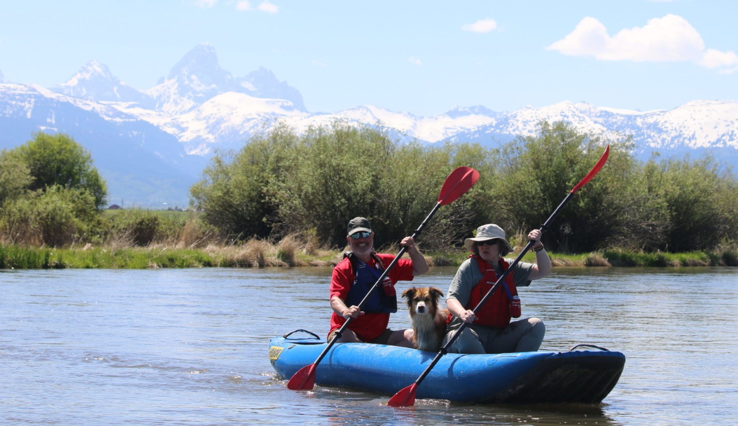 Two people with a dog kayaking on a river, mountains in the background.