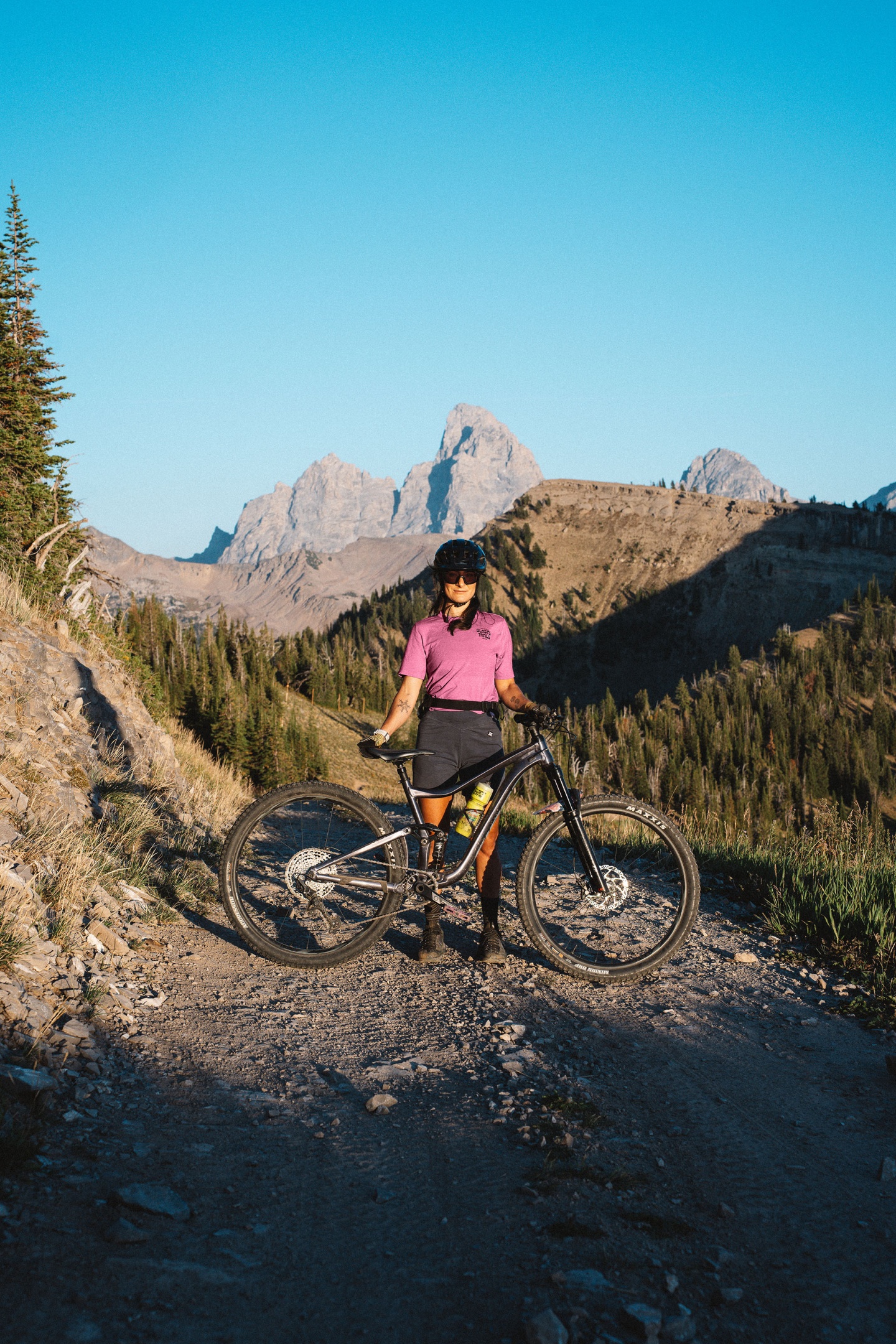 Cyclist on a mountain trail with distant peaks under clear blue sky.