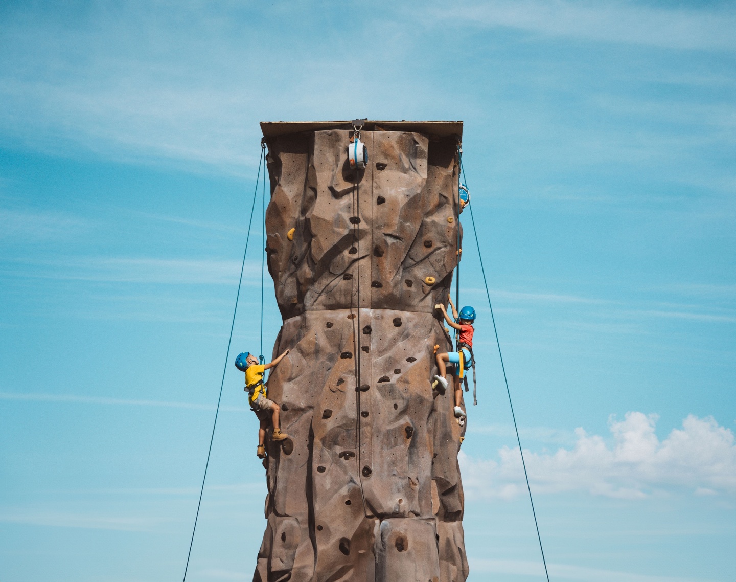 Climbers in helmets scale a tall outdoor rock wall against a blue sky.