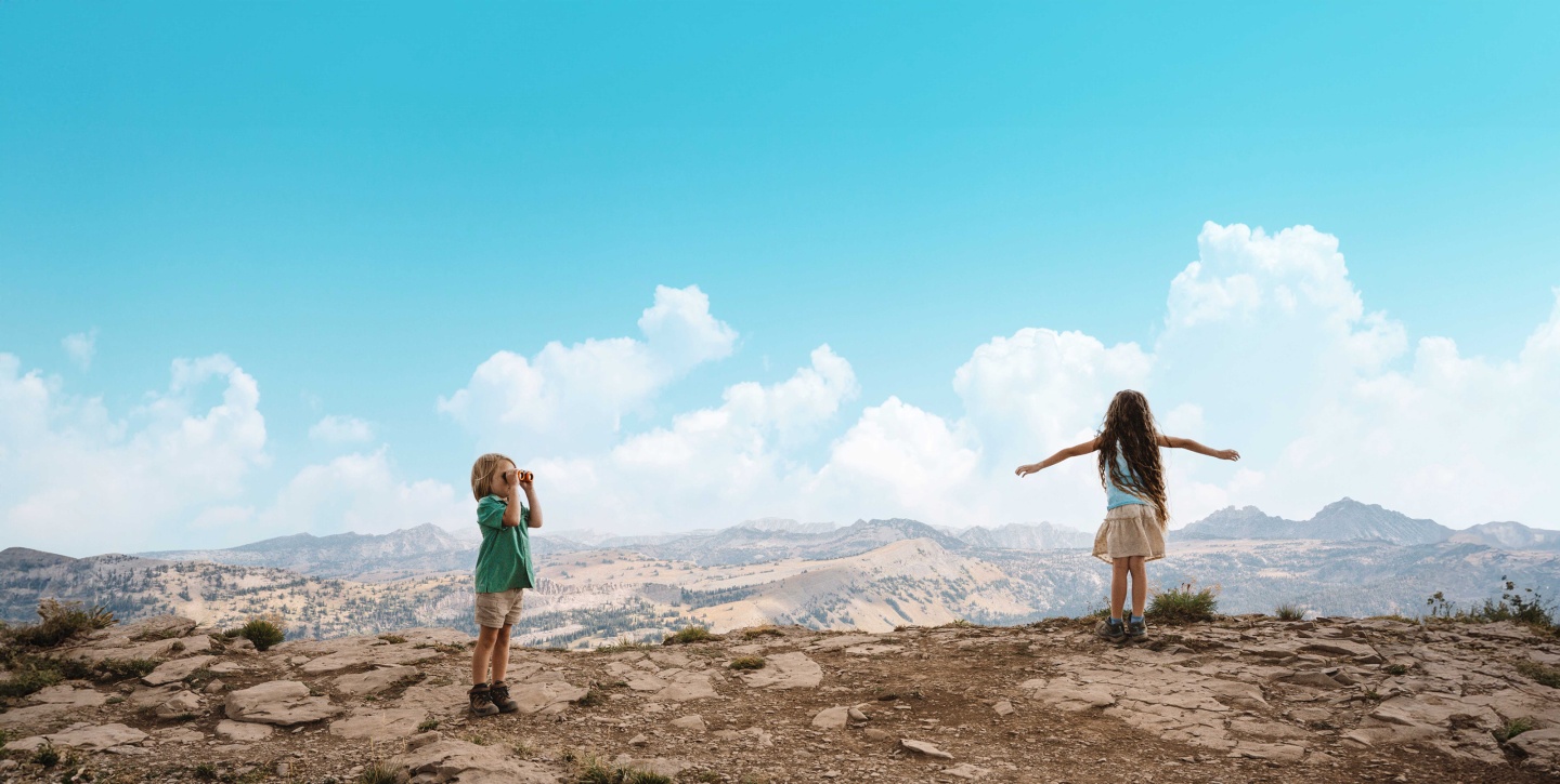 Children on a rocky hilltop, one with binoculars, clear blue sky.