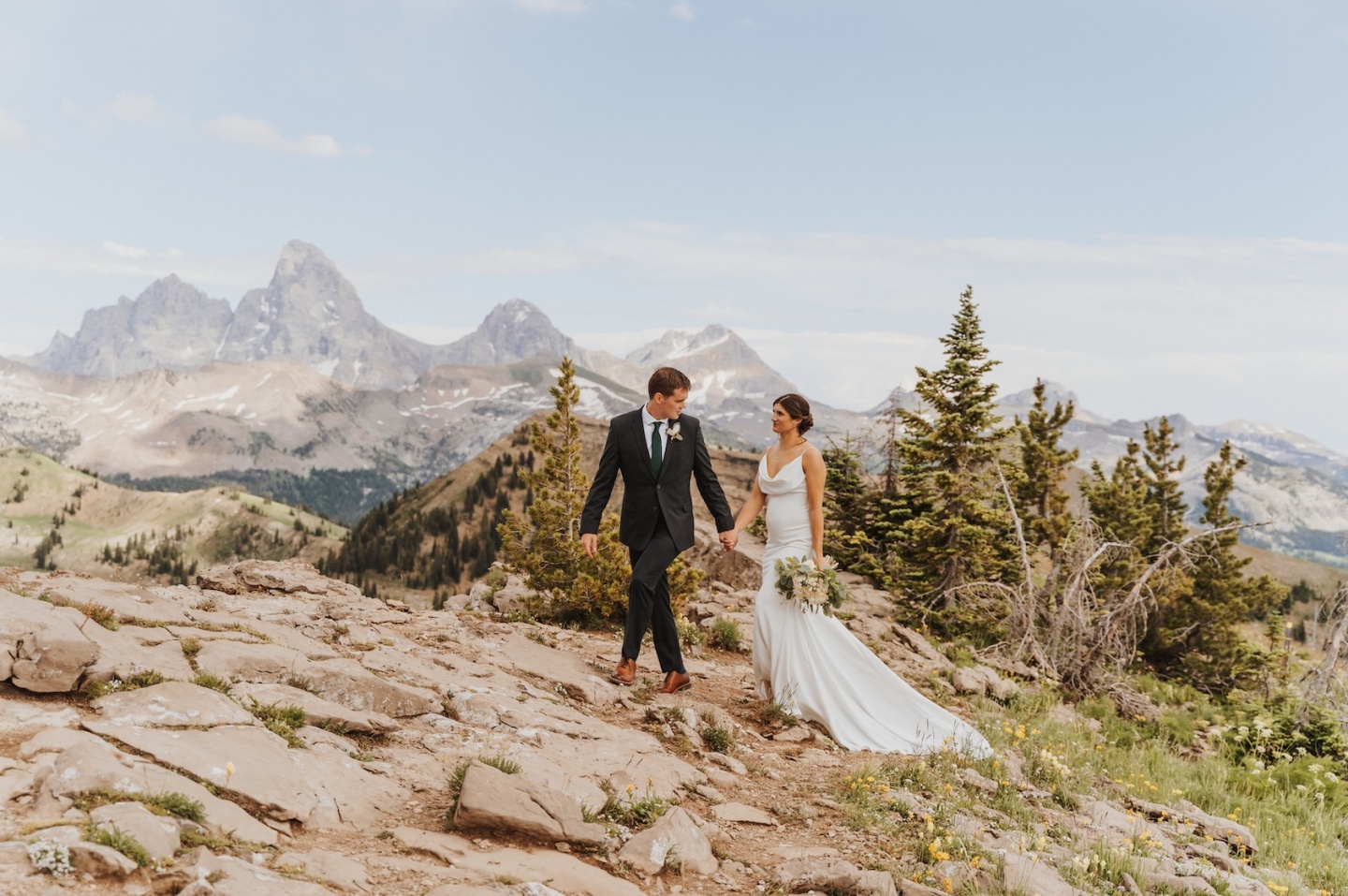 Couple in wedding attire walking on a rocky mountain path.