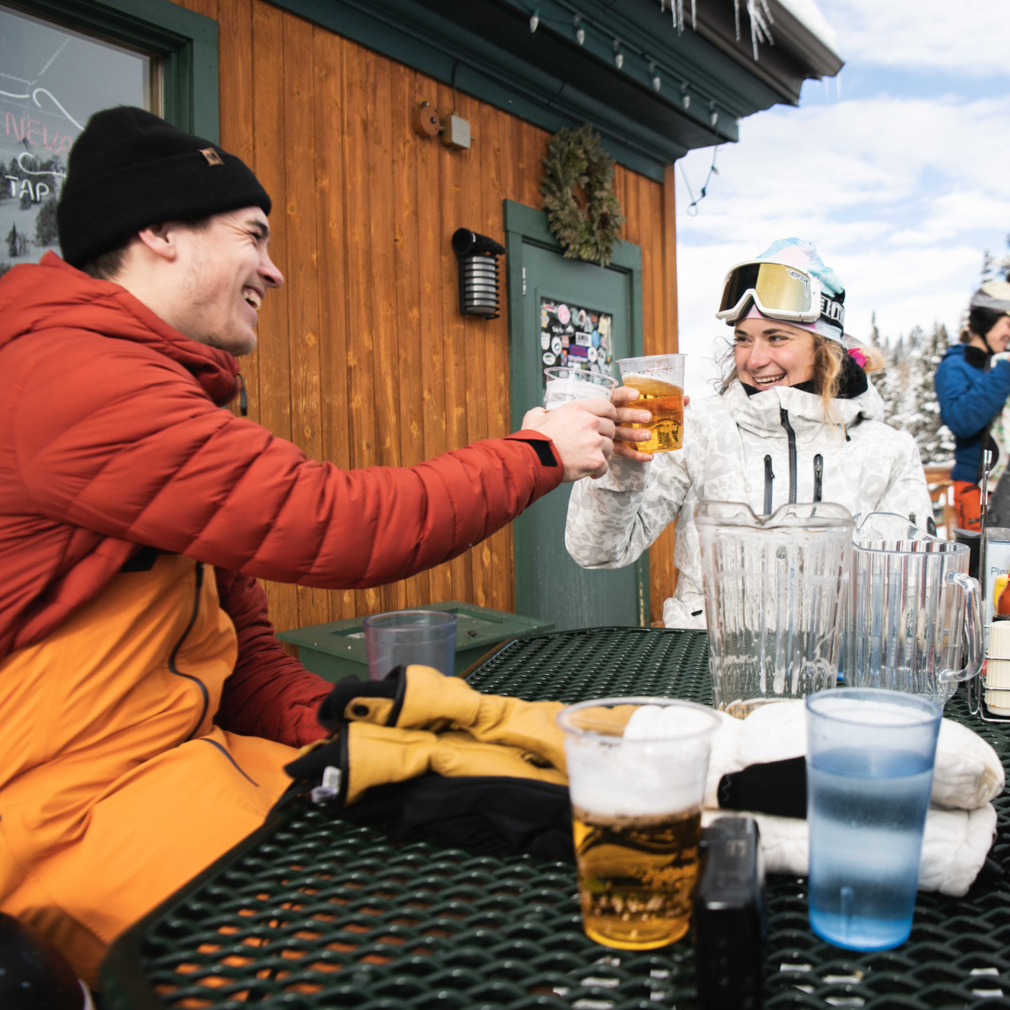 Two people in ski gear toast with beers at an outdoor table.