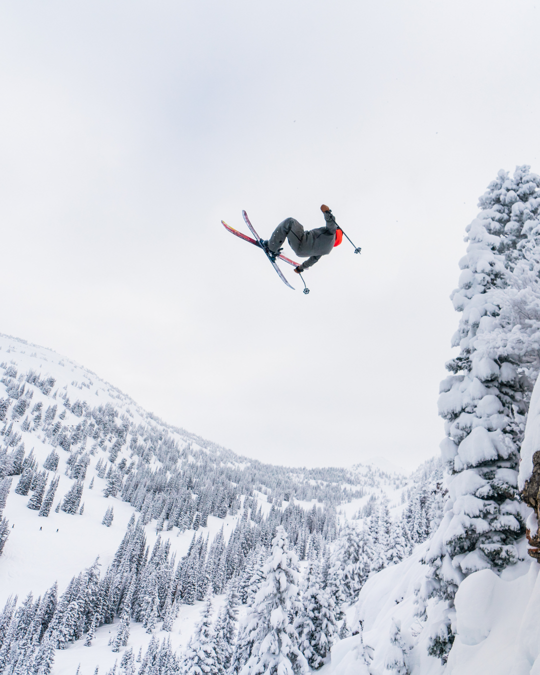 Skier performing a flip in snowy mountains, surrounded by trees.