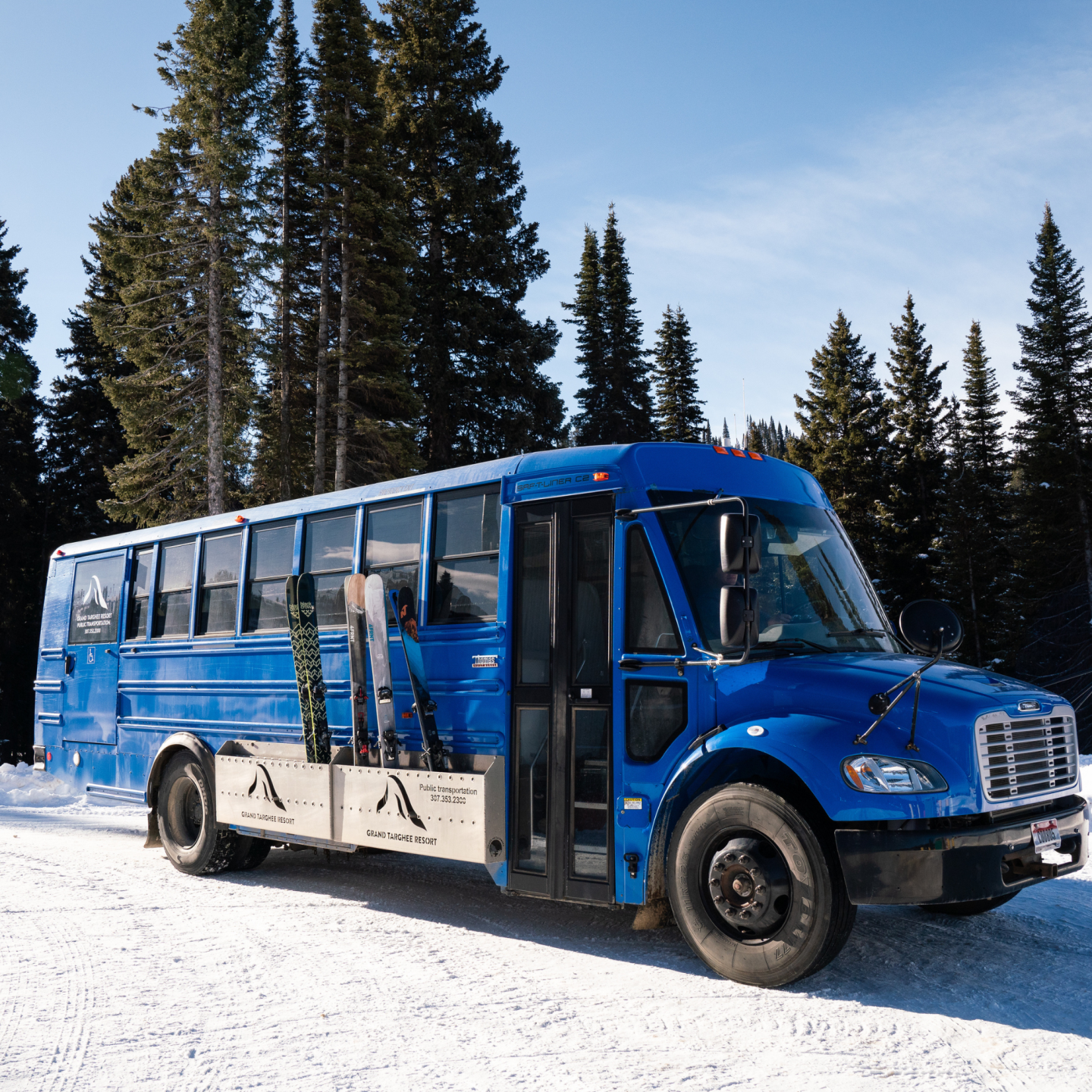 Blue ski bus parked on snowy road, surrounded by pine trees.
